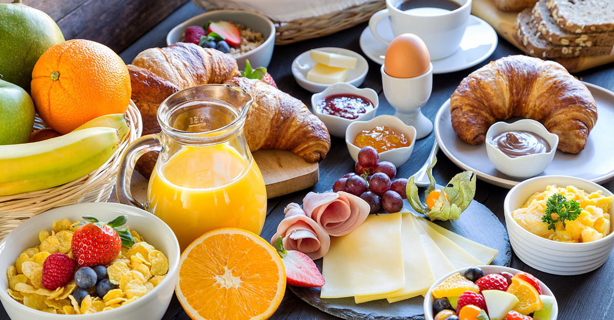 Colazione con croissant, uova strapazzate, salumi, formaggi, frutta, cereali, succo d'arancia e caffè.