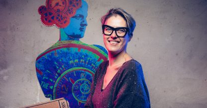 Una persona sorridente con gli occhiali si trova di fronte a un'opera d'arte colorata a tema meccanico ispirata agli assorbenti.