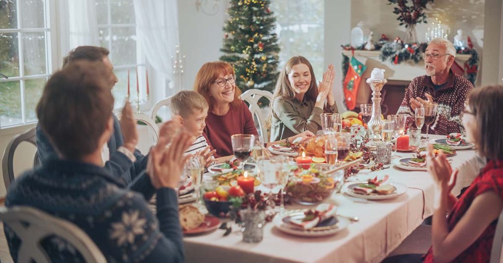 Un gruppo si gode un convivio attorno a un tavolo festivo, sorridendo e applaudendo con le decorazioni natalizie sullo sfondo.