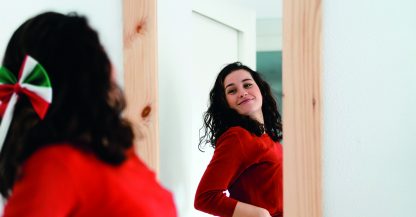 Giovane bella donna sicura di sé in maglione rosso che si guarda allo specchio orgogliosa di se stessa e che usa cosmetici